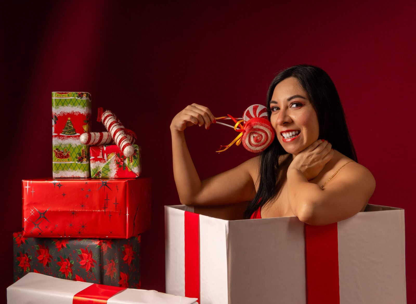 Joyful Latina woman with candy cane surrounded by Christmas gift boxes on a red backdrop.